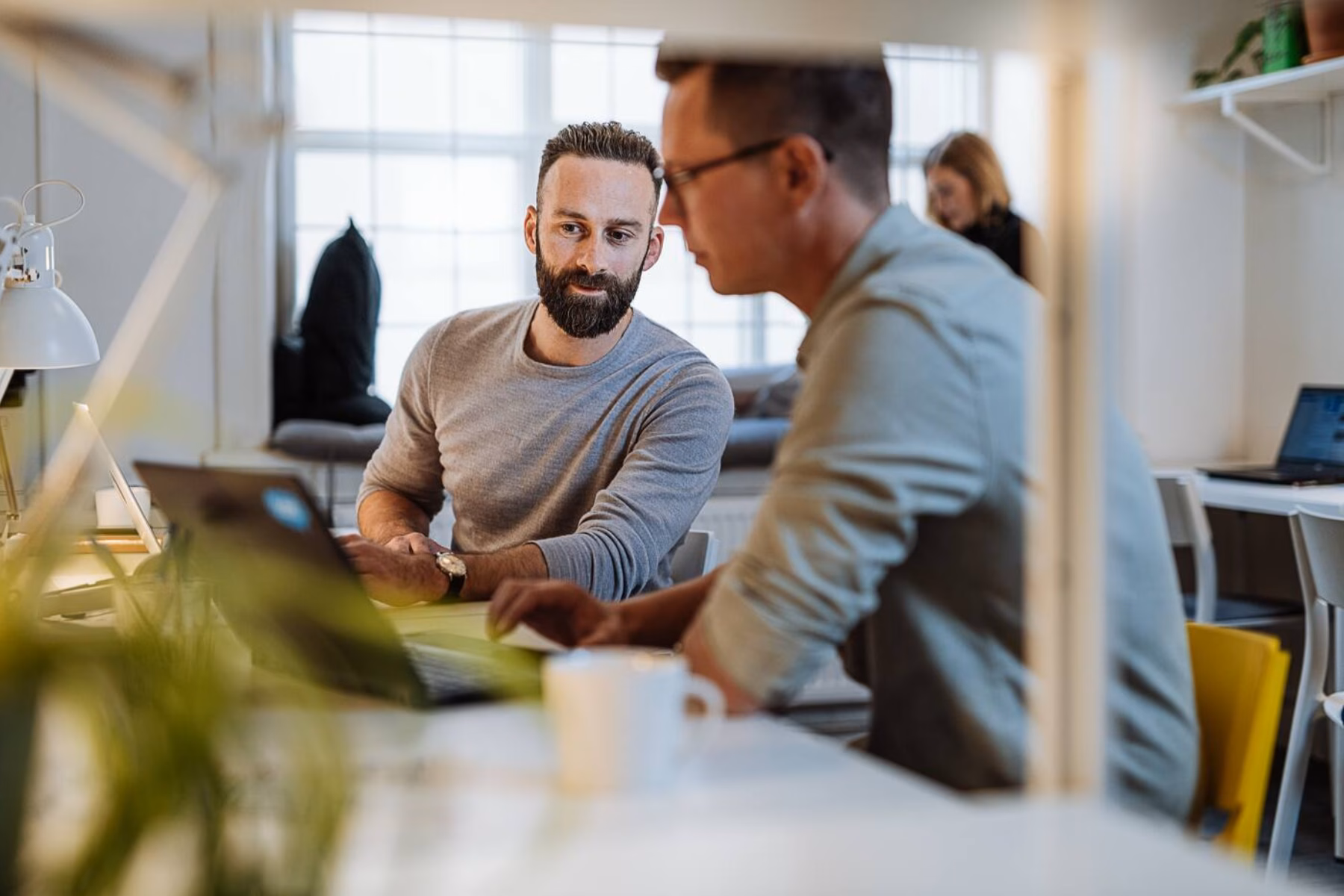 Two men are working in a shared office space, with one man showing the other something on his laptop.