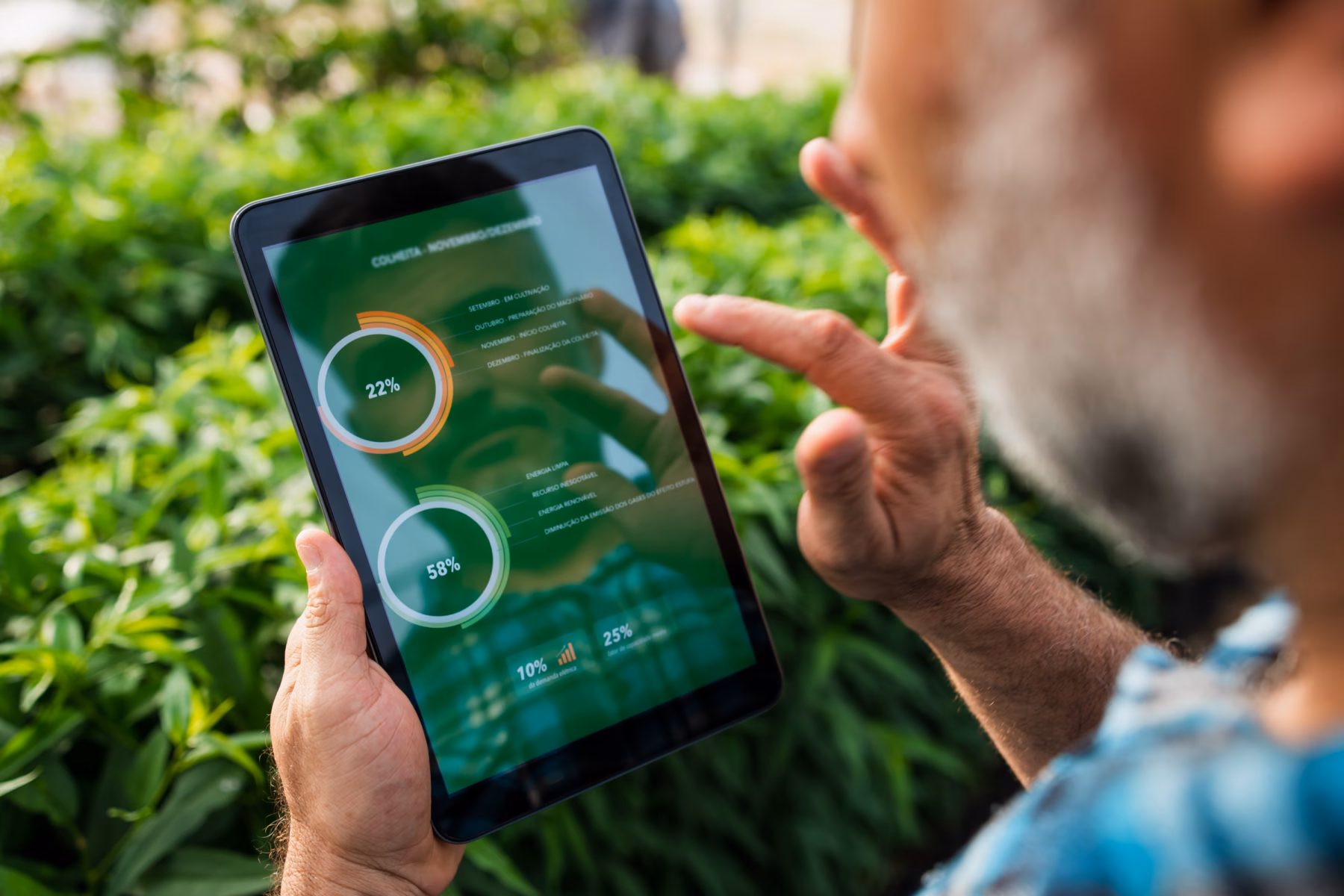 A man is in a green environment with bushes, looking at data on a tablet.