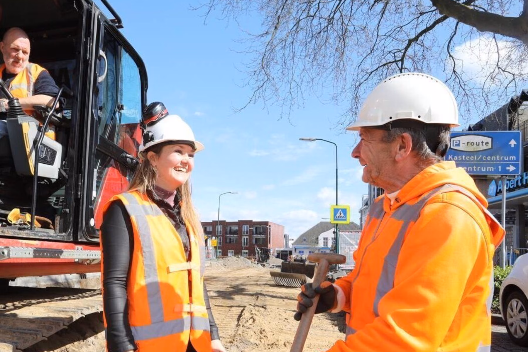 Workers at a construction site are wearing reflective vests and safety helmets, with one operating a excavator while a man and a woman stand in front, smiling and working. The man is wearing gloves and holding a shovel.