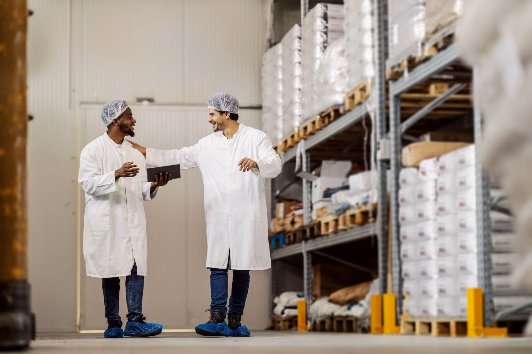Full-length shot of two food factory warehouse workers walking towards the camera, talking and collaborating, with one holding a tablet.