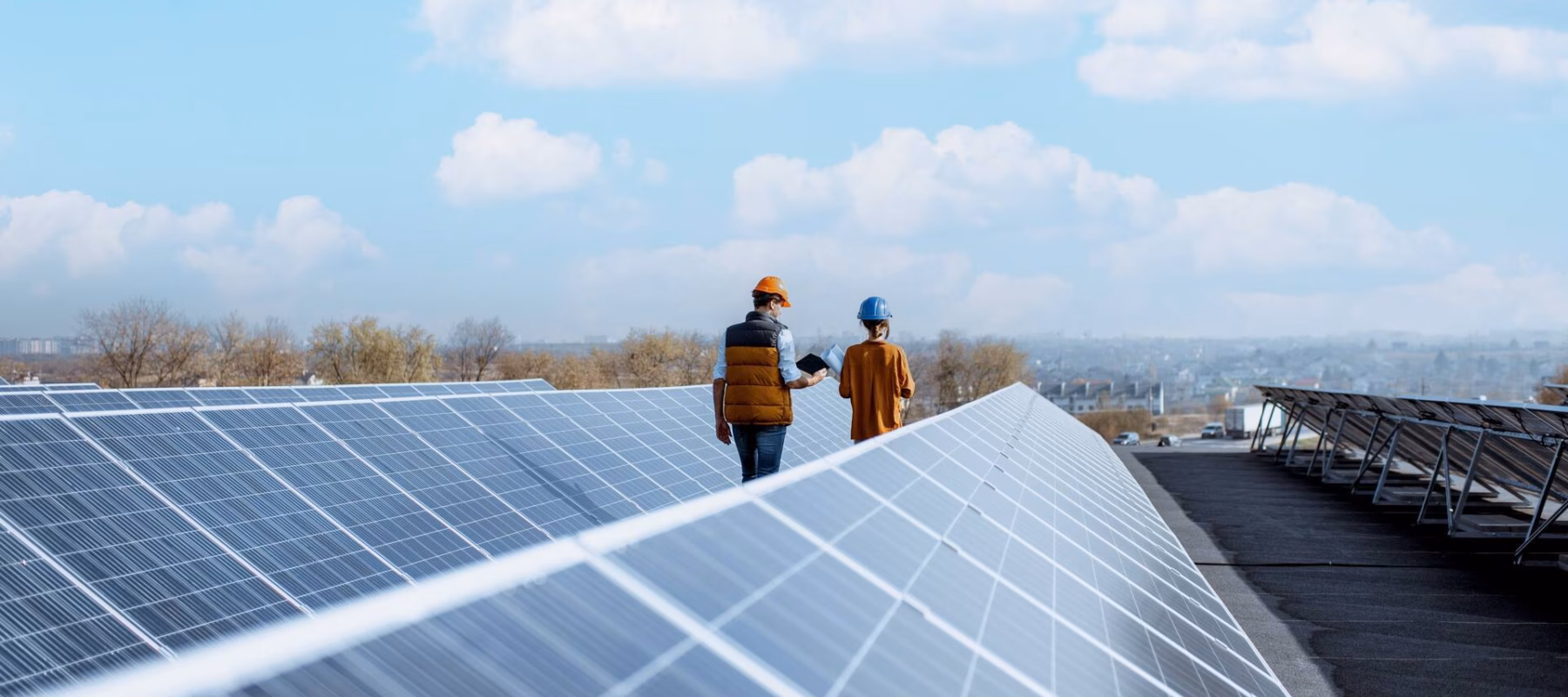 Men are inspecting solar panels on a roof under a light blue sky, wearing safety helmets.