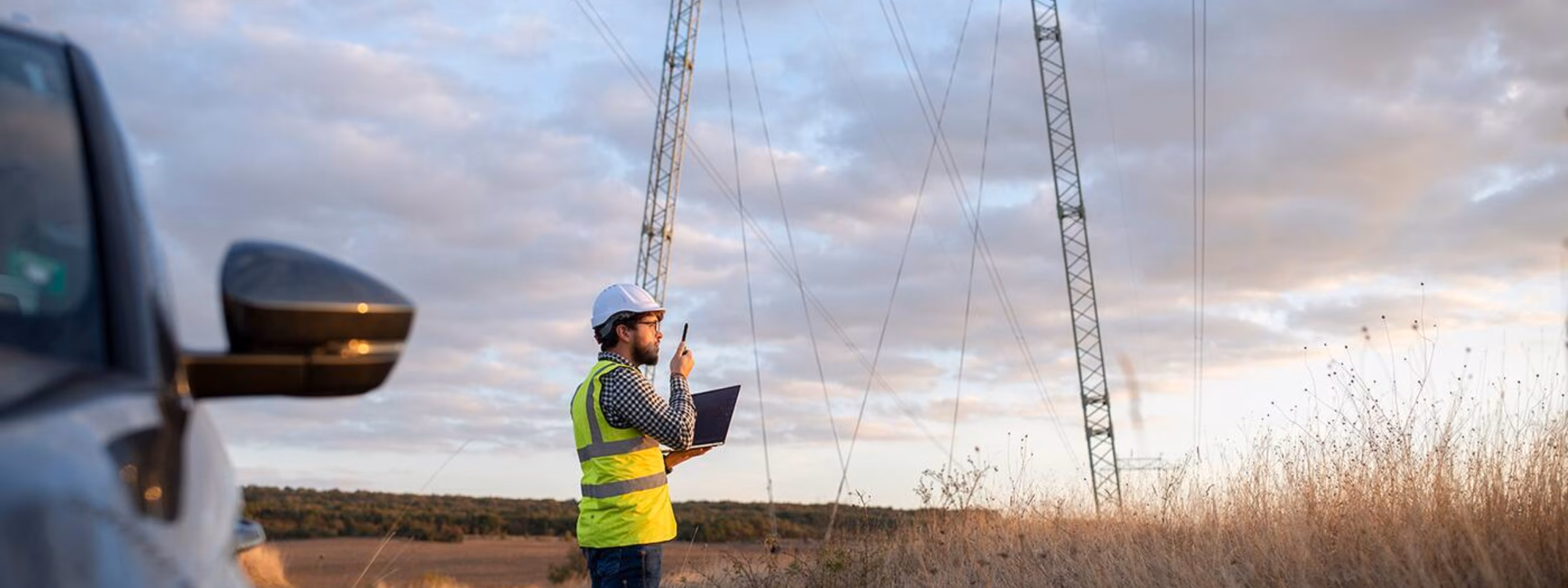 A person wearing a safety vest uses a tablet near high-voltage power lines during a field inspection in an open landscape.