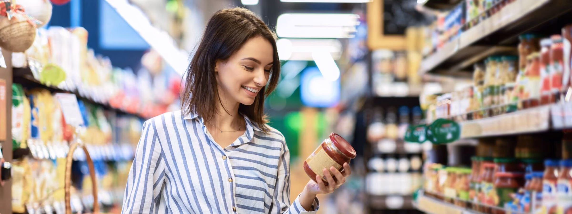 Een lachende vrouw loopt door de gangpaden van een supermarkt met een winkelwagen en bekijkt een glazen pot saus, terwijl ze gezonde producten kiest tussen de schappen.