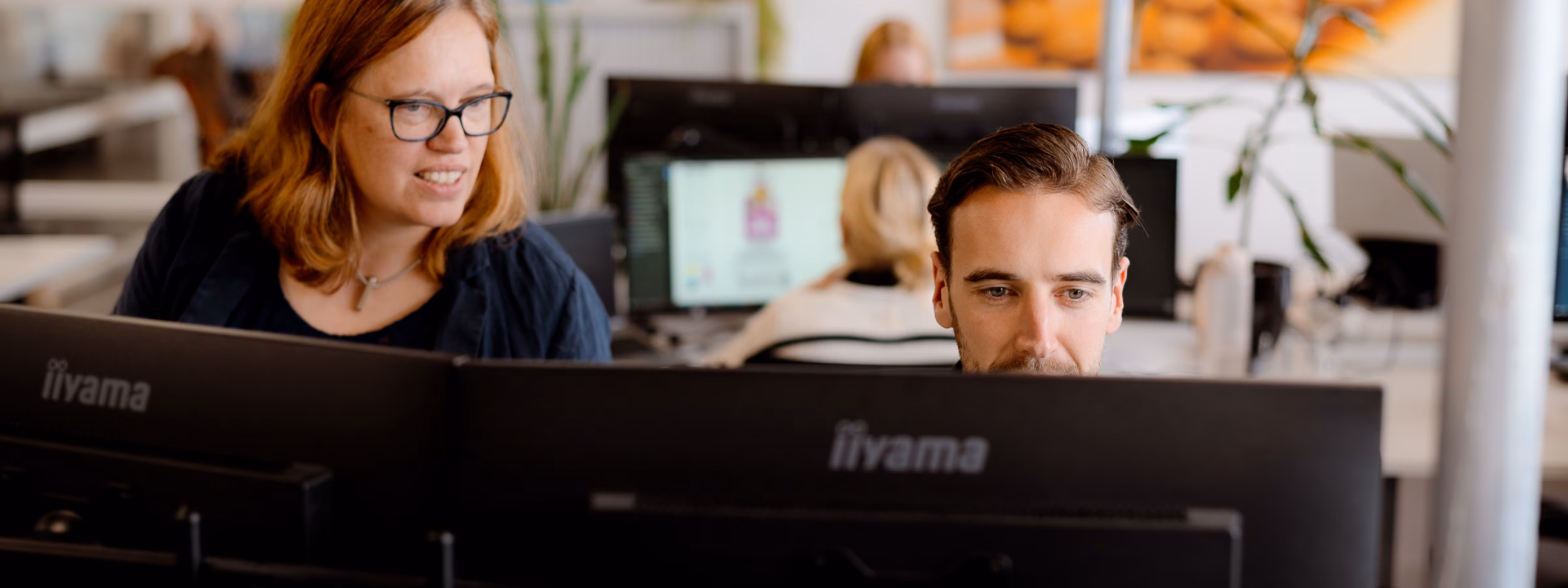 Two employees are working together behind multiple computer screens in a modern office environment.
