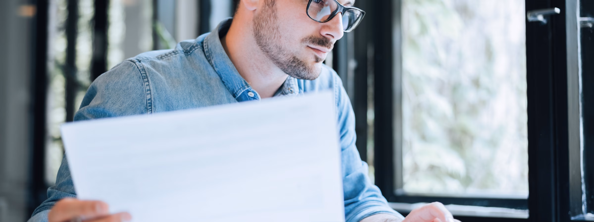 A man is typing and looking at his laptop while holding a paper in his hand, working at his desk.