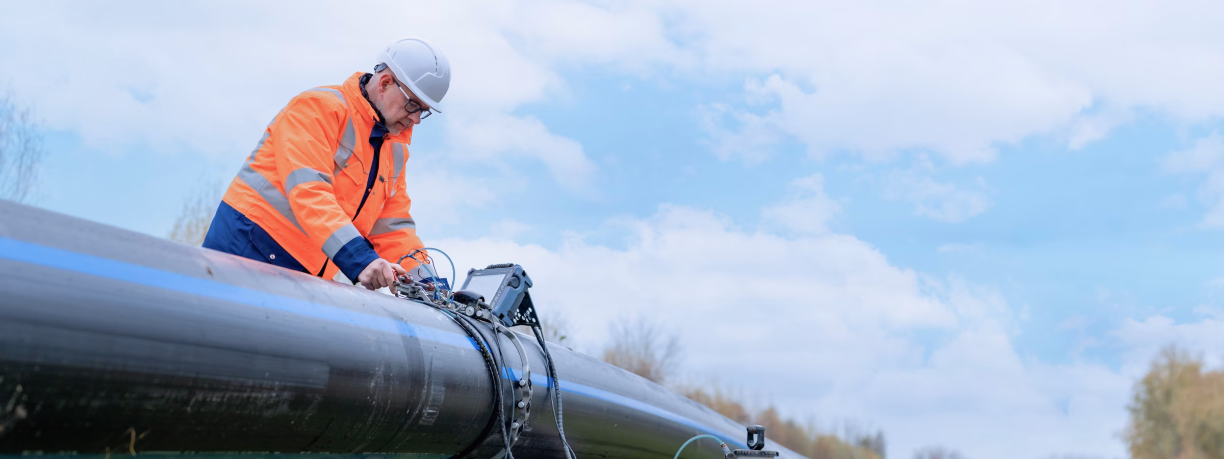 A man is inspecting a large pipe in a grassy field, wearing a reflective jacket and a safety helmet.