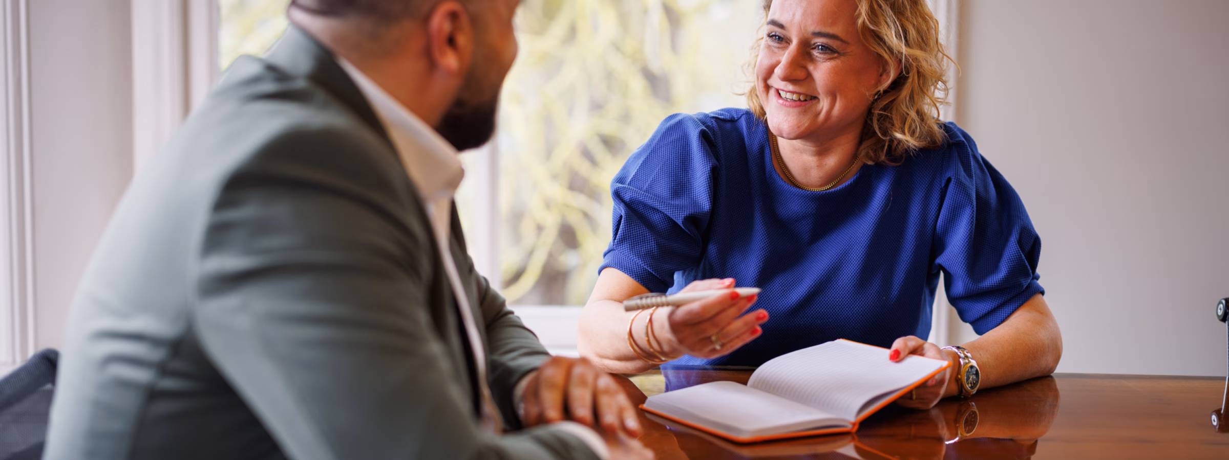 Man and woman in a meeting, with the woman wearing a blue top taking notes while smiling during the conversation.