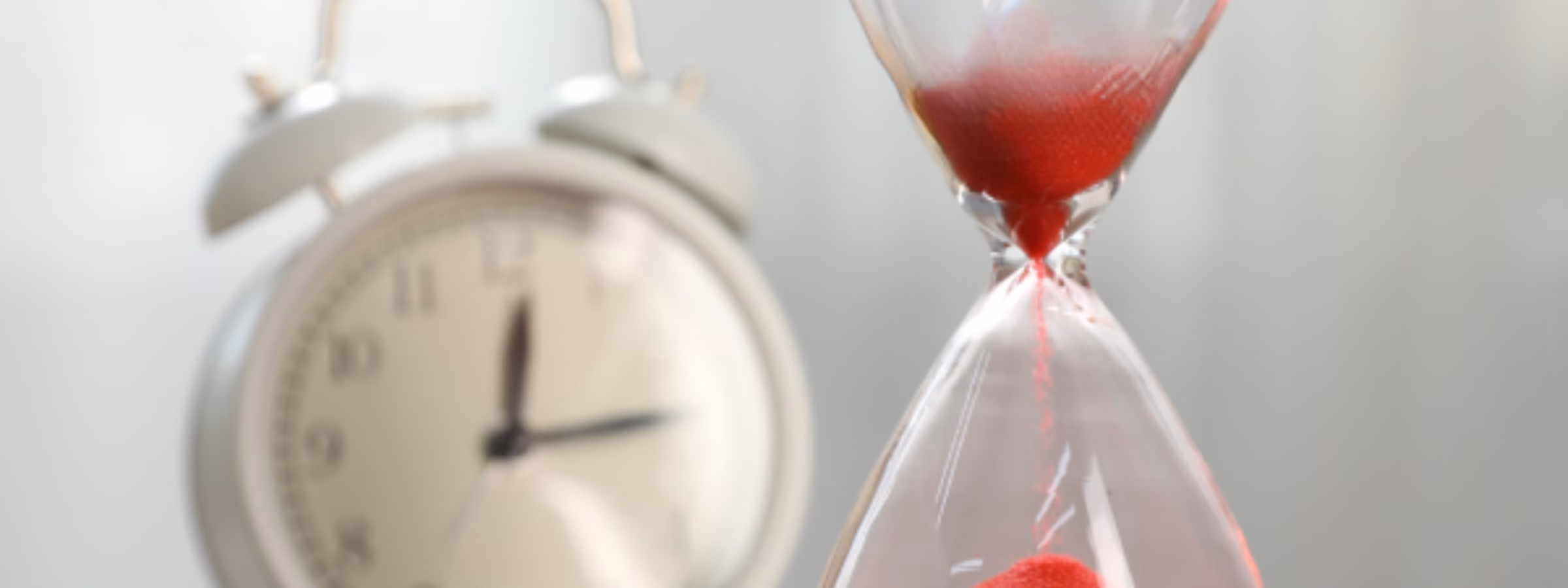 Im Vordergrund steht neben einem klassischen Wecker eine Sanduhr mit rotem Sand, die für schnelle Bearbeitungszeiten, effiziente Dienstleistungserbringung, Terminmanagement, zeitkritische Projekte und reaktionsschnellen Support bei Prüf-, Inspektions-, Zertifizierungs- und Konformitätsaktivitäten steht.