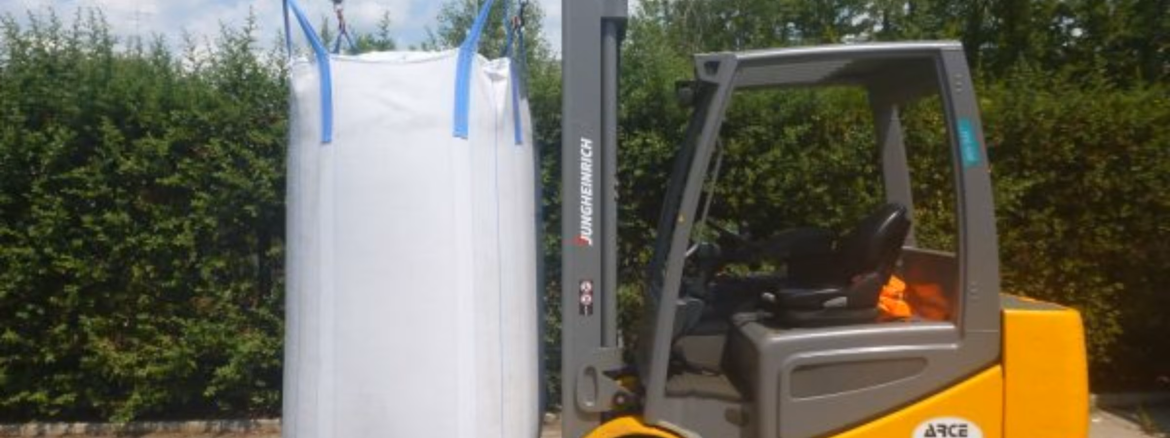 Ein Gabelstapler transportiert einen großen industriellen Schüttgut-Sack mithilfe eines speziellen Hubaufsatzes in einem Logistikbereich im Freien, wobei der Schwerpunkt auf sicherem Materialumschlag, Schüttguttransport, Lagereffizienz, Qualitätssicherung und industriellem Lieferkettenmanagement liegt