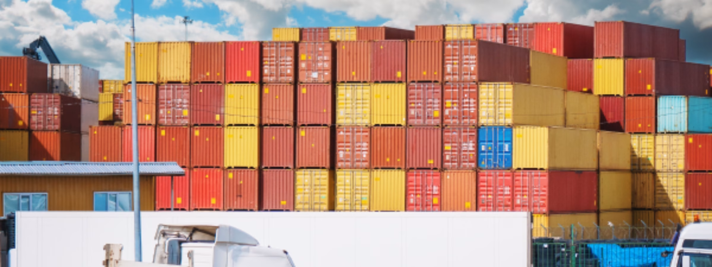 Vor gestapelten Versandcontainern in einem geschäftigen Logistikterminal sind Lastkraftwagen im Einsatz, was die Bedeutung von globalem Lieferkettenmanagement, Gütertransport, internationalem Handel, Lagerhaltung und effizienten Distributionsdienstleistungen innerhalb eines modernen Logistiknetzwerks verdeutlicht.