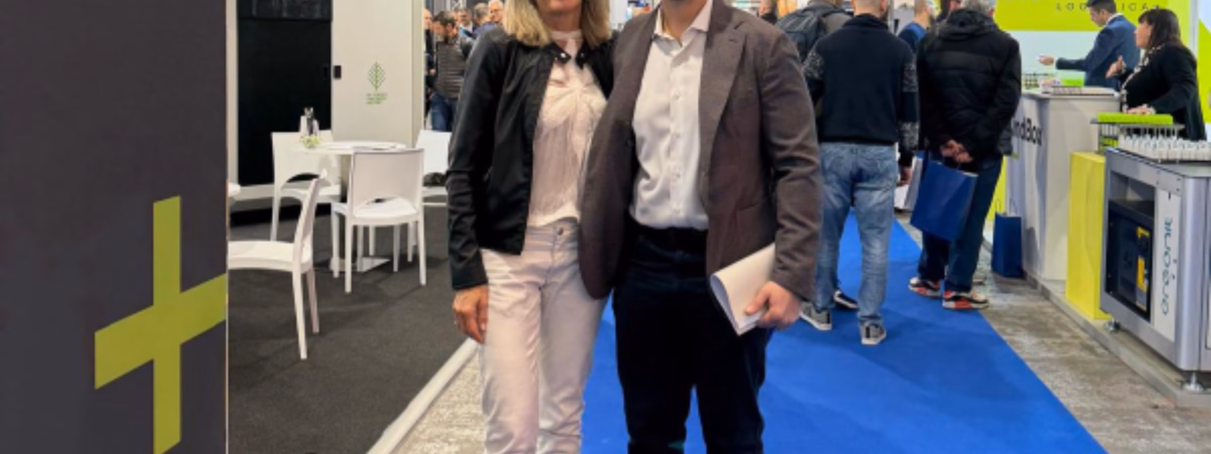 Zwei Fachleute stehen gemeinsam auf der Ausstellungsfläche einer Fachmesse, umgeben von Messeständen der Branche und Besuchern. Im Vordergrund stehen dabei geschäftliches Networking, branchenübergreifende Zusammenarbeit, Wissensaustausch sowie der Austausch über Prüf-, Inspektions-, Zertifizierungs- und Konformitätsdienstleistungen.
