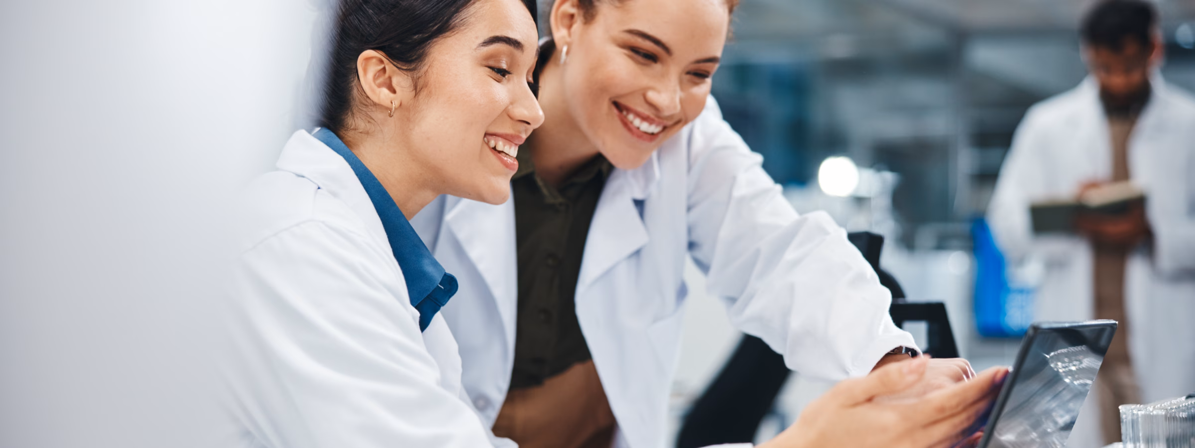 Zwei Frauen in Laborkitteln lächeln, während sie in einem Labor auf einen Laptop blicken; in der Nähe sind wissenschaftliche Geräte zu sehen.