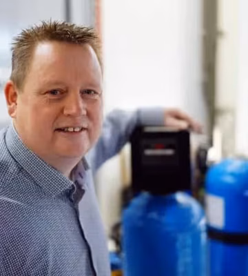 Die Person steht neben Geräten zur Wasseraufbereitung oder Wasseranalyse in einer Labor- oder technischen Arbeitsumgebung.