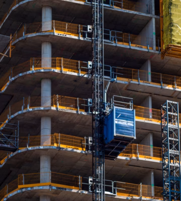 Plusieurs étages d’un immeuble de grande hauteur en béton sont visibles pendant la phase de gros œuvre. Un ascenseur de chantier longe la façade pour assurer le transport vertical des matériaux et du personnel. Aux étages supérieurs, une charge de chantier emballée est hissée, tandis que des barrières de sécurité ont été installées le long des bords des planchers.