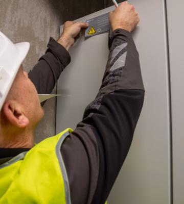 Un employé portant un équipement de protection individuelle appose une étiquette d'identification ou d'avertissement sur une armoire électrique. Un symbole d'avertissement indiquant la présence de tension électrique est visible sur l'armoire. L'illustration montre des travaux dans les domaines de l'électrotechnique, de la gestion des installations et de la sécurité.