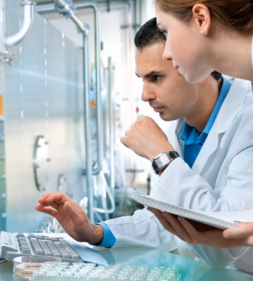 A man and a woman wearing white coats are reviewing information on a computer in a laboratory setting, with the woman holding a notebook.