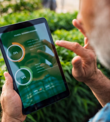 A man is in a green environment with bushes, looking at data on a tablet.