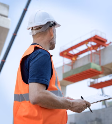 A person wearing a safety helmet and an orange safety vest at a construction site, with a building structure visible in the background.