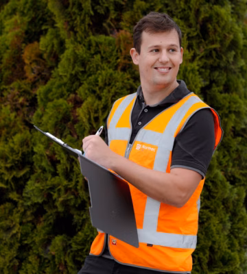 A smiling man is wearing a safety vest and holding a pen and notepad.