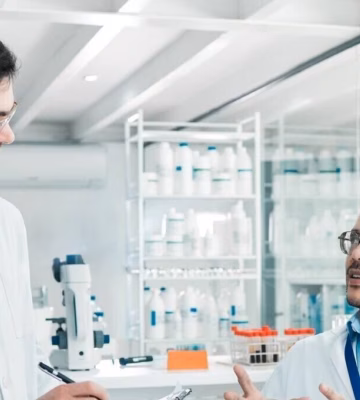 A group of scientists in a lab are wearing lab coats and discussing together while taking notes on a clipboard.