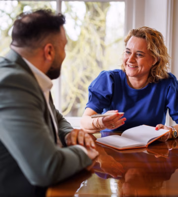 Man and woman in a meeting, with the woman wearing a blue top taking notes while smiling during the conversation.