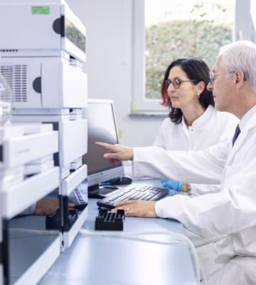 Two laboratory technicians are reviewing analytical data at a computer workstation located in close proximity to state-of-the-art laboratory equipment. This image illustrates scientific collaboration, research, quality control, and accuracy testing in a modern laboratory.