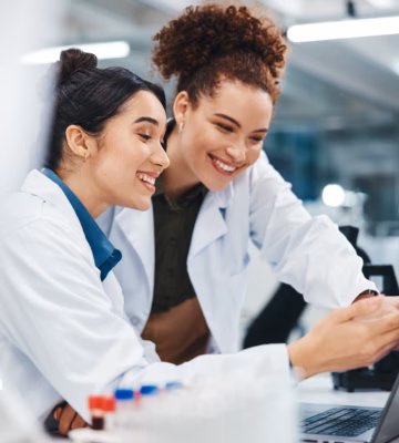 Two women in lab coats are smiling as they look at a laptop in a lab; scientific equipment can be seen nearby.