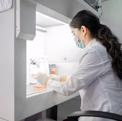A woman is testing samples in petri dishes in a laboratory, wearing a lab coat and a face mask.