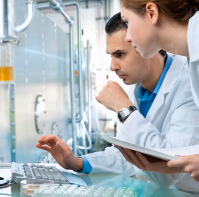 Un homme et une femme vêtus de blouses blanches examinent des informations sur un ordinateur dans un laboratoire, la femme tenant un cahier.