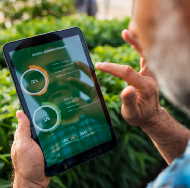 A man is in a green environment with bushes, looking at data on a tablet.