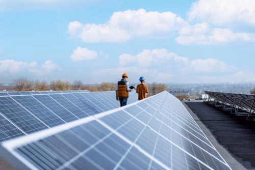 Men are inspecting solar panels on a roof under a light blue sky, wearing safety helmets.