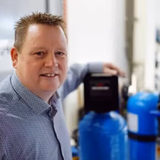 A person is standing next to water treatment or water analysis equipment in a laboratory or technical work environment.