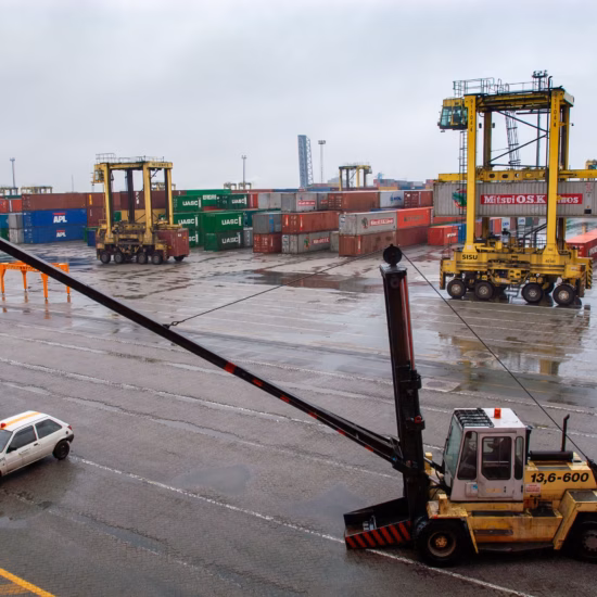 Op een uitgestrekt containerterrein staan rijen zeecontainers opgesteld terwijl meerdere portaalhefwagens containers verplaatsen. Het natte wegdek en de grijze lucht wijzen op regenachtig weer. Op de voorgrond staat een reach stacker, met daarachter de containeropslag en overslagactiviteiten in de haven.