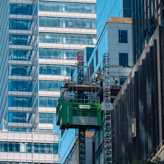Un chantier est en cours entre de grands immeubles de bureaux aux façades vitrées. Un ascenseur de chantier écologique permet d'accéder à une plate-forme de travail le long de la façade, tandis que les ouvriers effectuent des travaux en hauteur. L'image montre la combinaison de gratte-ciel modernes, de travaux de façade et de transport vertical sur un chantier urbain.