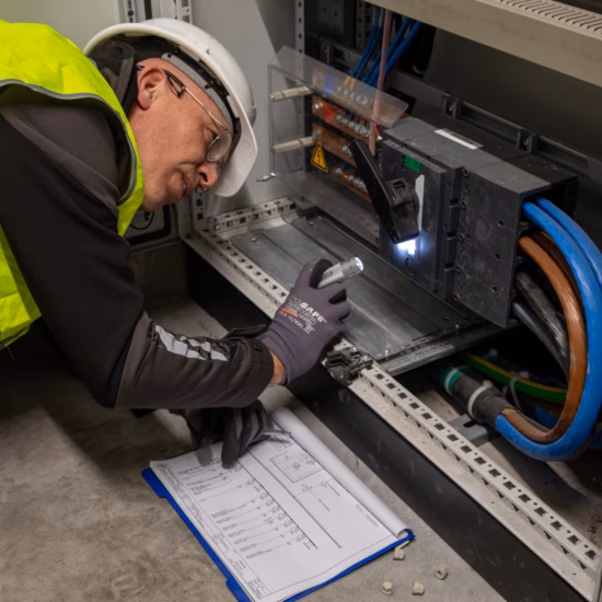 Een technicus draagt persoonlijke beschermingsmiddelen en voert een inspectie uit in een geopende elektrische verdeelkast. Met behulp van een zaklamp en technische documentatie controleert de technicus de bekabeling, aansluitingen en componenten van de elektrische installatie.