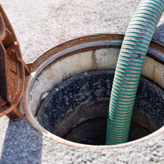 Un regard d'égout ouvert dans une chaussée pavée, avec un tuyau flexible vert placé à l'intérieur du regard. L'image montre des travaux sur le réseau d'égouts, tels que l'inspection, le nettoyage, la vidange ou l'entretien des infrastructures souterraines.