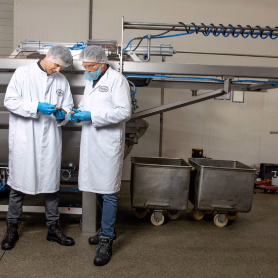 Twee mannen in witte steriele labjassen en met haarnetjes overleggen in een fabriekshal, terwijl ze productie- of kwaliteitsprocessen bespreken.