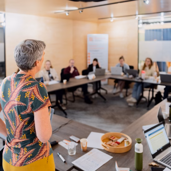 Deelnemers nemen deel aan een bijeenkomst of training in een vergaderruimte, terwijl een spreker voor de groep presenteert.