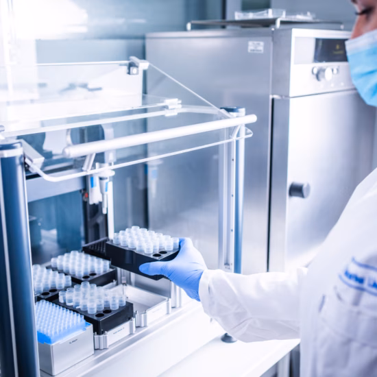Een vrouw in een witte labjas en mondkapje haalt meerdere reageerbuisjes uit een rek in een laboratorium, geconcentreerd werkend aan een experiment.