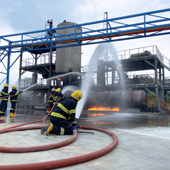 Firefighters in full firefighting gear are extinguishing an industrial fire using water.
