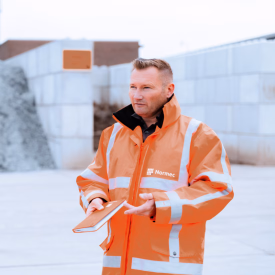 Men wearing orange coats are conversing on a construction site, with one man holding a notebook.