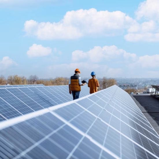Men are inspecting solar panels on a roof under a light blue sky, wearing safety helmets.