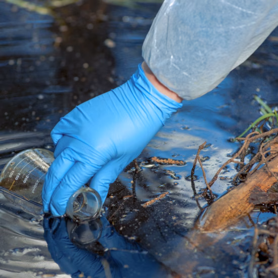 Medewerker met blauwe handschoen neemt een watermonster uit oppervlaktewater met een glazen monsterfles.