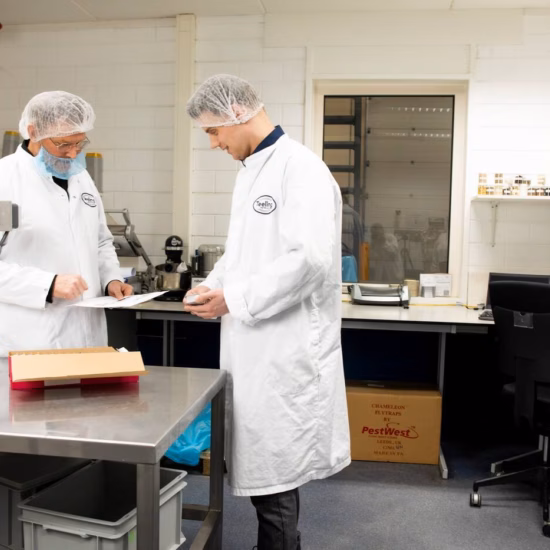 Men in white coats and hairnets are inspecting a box, discussing while one man looks at a sheet of paper.