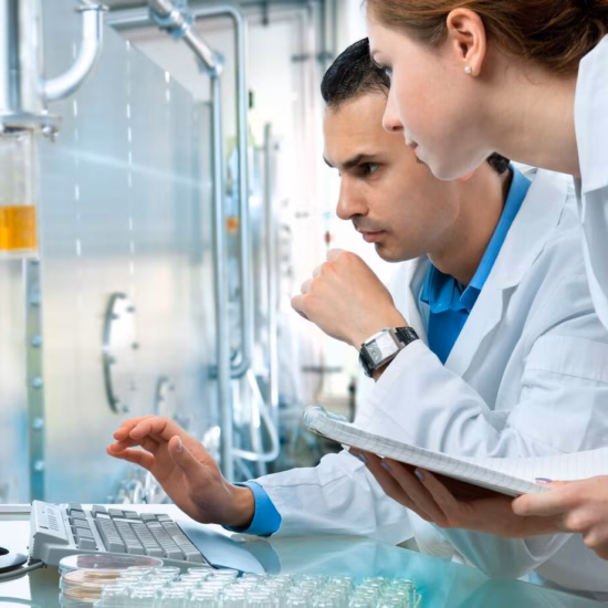 A man and a woman wearing white coats are reviewing information on a computer in a laboratory setting, with the woman holding a notebook.