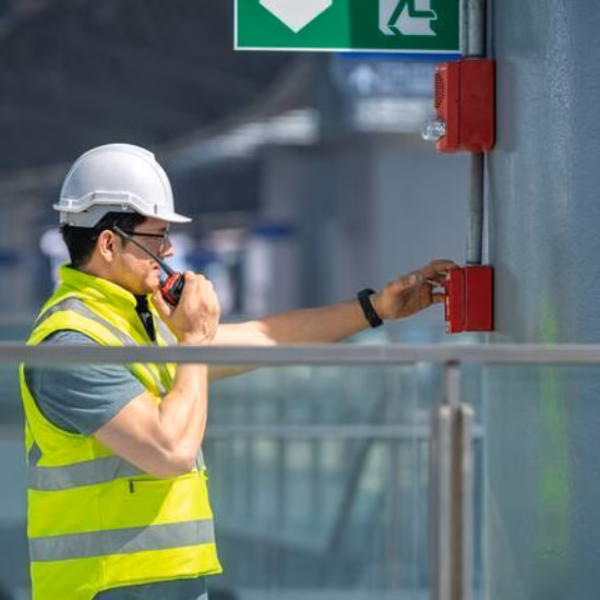 An engineer wearing a safety uniform and helmet is checking the fire alarm emergency system in an industrial factory, with an exit door visible.