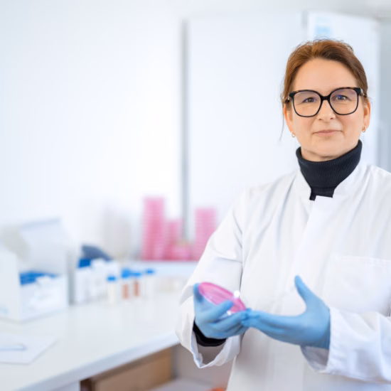 A woman is looking at the camera in a laboratory while holding a petri dish, wearing a lab coat and blue sterile laboratory gloves.