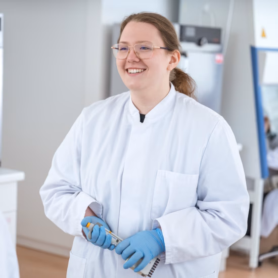 A laboratory scientist is smiling at a colleague in a laboratory, holding testing equipment, and wearing a lab coat and blue sterile laboratory gloves.