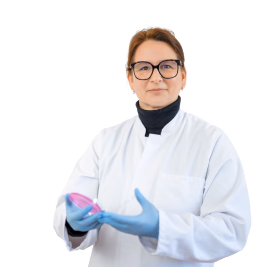 A woman is looking at the camera in a laboratory while holding a petri dish, wearing a lab coat and blue sterile laboratory gloves.