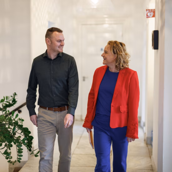 Man and woman walking through an office hallway together while smiling and discussing work.